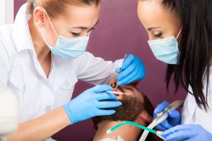 Two oral hygienists with masks and gloves performing professional teeth cleaning on a patient.
