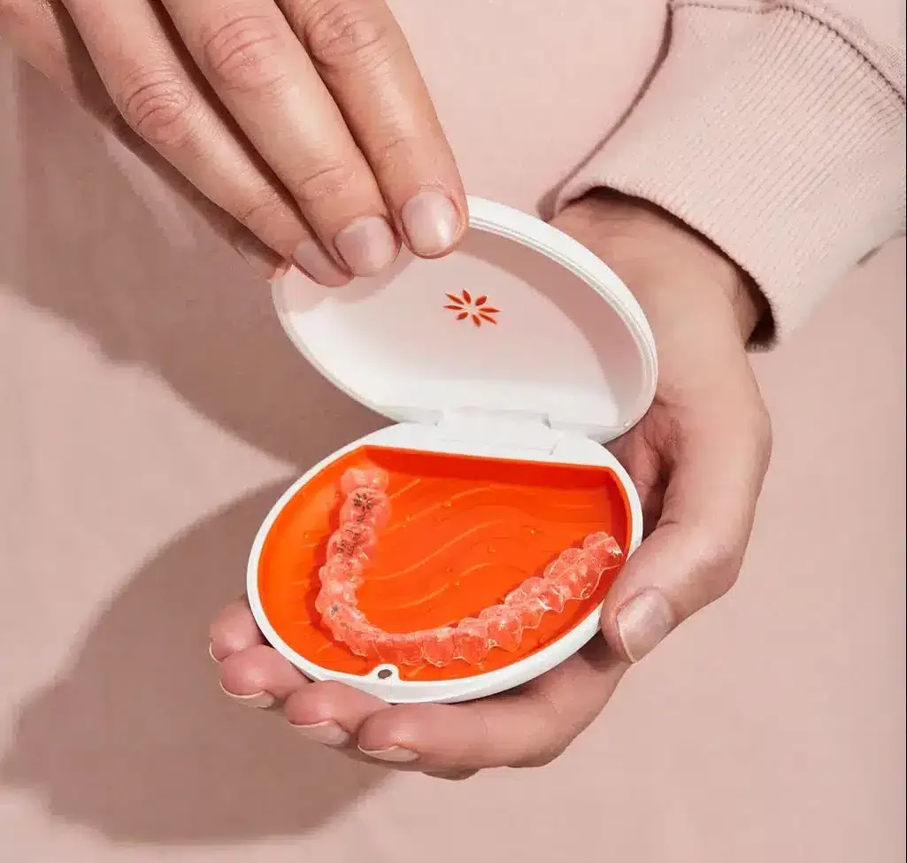 Oseba drži bel etui z oranžno notranjostjo, v katerem je nevidni zobni aparat Invisalign.