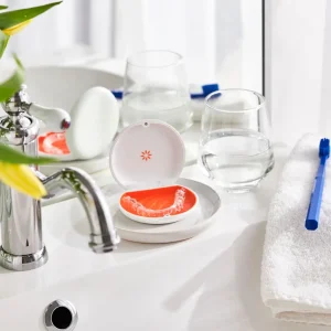 Prozorna zobna opornica Invisalign v belem in oranžnem etuiju na kopalniškem pultu ob kozarcu vode in zobni ščetki.