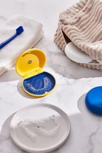 Nevidni zobni aparat Invisalign v rumeno-modrem etuiju na kopalni pultu z brisačo in zobno ščetko.