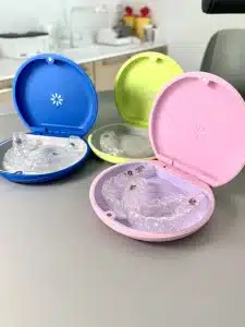 Barviti etuiji za Invisalign opornice (modri, rožnati, zeleni) z nevidnimi zobnimi opornicami na zobozdravniški mizi.
