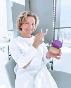 Nasmejana ženska v beli obleki drži Invisalign opornico in vijoličen etui v zobozdravniški ordinaciji.